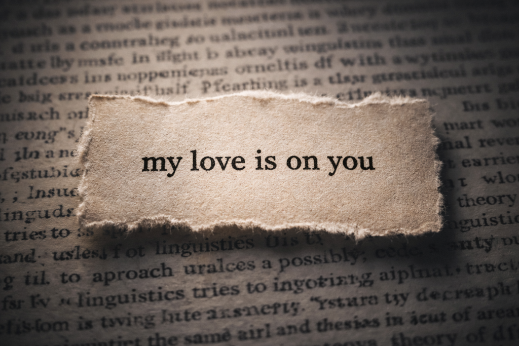 A close-up of a torn piece of aged paper with the phrase 'my love is on you' printed in serif typewriter font, resting on a page of a vintage book. An example of Scots' Gaelic linguistic nuance used to illustrate structural depth in micro fiction.